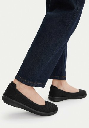 Czarne buty wsuwane z siateczki noszone z ciemnoniebieskimi jeansami o prostych nogawkach na jednolitym tle.