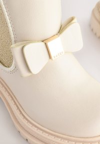 Bottine beige clair en matière synthétique texturée, dotée d'un nœud décoratif et d'un accent carré doré. Semelle robuste, finition lisse.