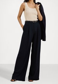 Beige kabelstickad linne, marinblå vida byxor med plisseringar, silverspänne bälte och en mörk blazer hängande över axeln, med öppna tåklackar.