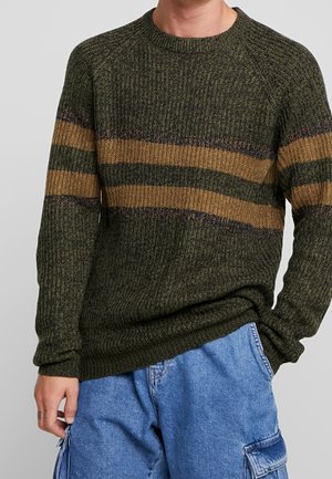 Mann trägt einen gerippten grünen Pullover mit braunen und dunkelgrünen horizontalen Streifen sowie blaue Cargohosen, die Hand in der Tasche.