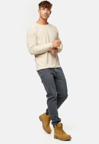 Beige Strickpullover mit langen Ärmeln, graue Jeans und hellbraune Schnürstiefel. Die Stiefel besitzen Metallösen und robuste Sohlen.
