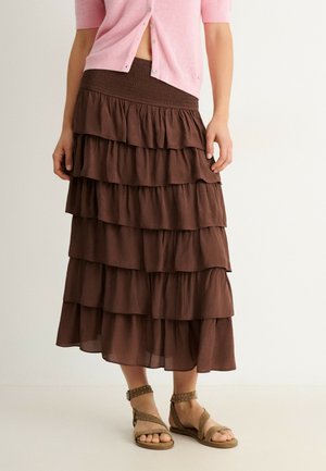 Femme portant une jupe midi marron à volants, un cardigan à manches courtes rose boutonné et des sandales beige cloutées avec bride à la cheville, debout devant un fond uni.