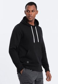 Sudadera negra de tela suave, con un bolsillo frontal, cordones blancos y puños acanalados. El modelo que la lleva puesta está de pie frente a un fondo neutro.