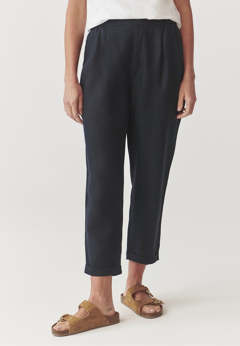 Personne portant un pantalon noir court et des sandales marron à bouts ouverts avec des brides à double boucle, debout devant un fond neutre.