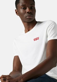 Mand iført en hvid Levi's t-shirt med et rødt logo på brystet, siddende med sammenfoldede hænder og kigger let til siden.