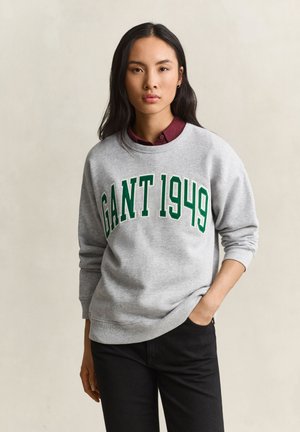 Jeune femme aux longs cheveux noirs portant un sweat-shirt gris avec "GANT 1949" en lettres vertes et un pantalon noir, une main dans la poche.