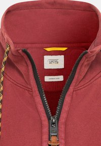 Sweat-shirt rouge teinture en vêtement avec un col haut, une fermeture éclair et une doublure texturée noire contrastante. Comprend un curseur de fermeture éclair en cuir marron.