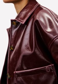 Bordeaux leren jas met een grote kraag, voor knopen en een patchzak met een logo. De stof heeft een glanzende, gestructureerde afwerking.