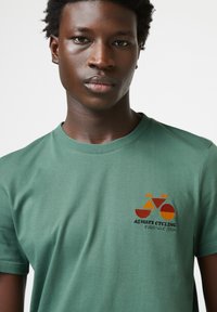 Grünes Baumwoll-T-Shirt mit einem bunten Fahrrad-Graphic und dem aufgenähten Text „ALWAYS CYCLING“ auf der Vorderseite. Kurze Ärmel und Rundhalsausschnitt.