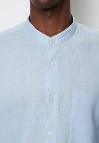 Vaaleansininen pellavapaita, jossa on pystykaulus, viisi nappia ja vasemmassa rinnassa läpällinen tasku. Teksturoitu, hengittävä kangas.