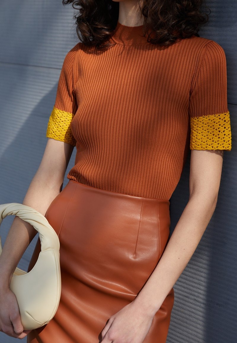 Ribbed rustinvärgad topp med gula virkade ärmar, i kombination med en figursydd brun läderkjol. Bär en handhållen krämfärgad, strukturerad handväska.