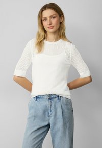 Witte gebreide trui met korte mouwen, voorzien van een zigzagpatroon. Draagt in combinatie met lichtblauwe high-waisted jeans. Eenvoudige ronde halslijn.