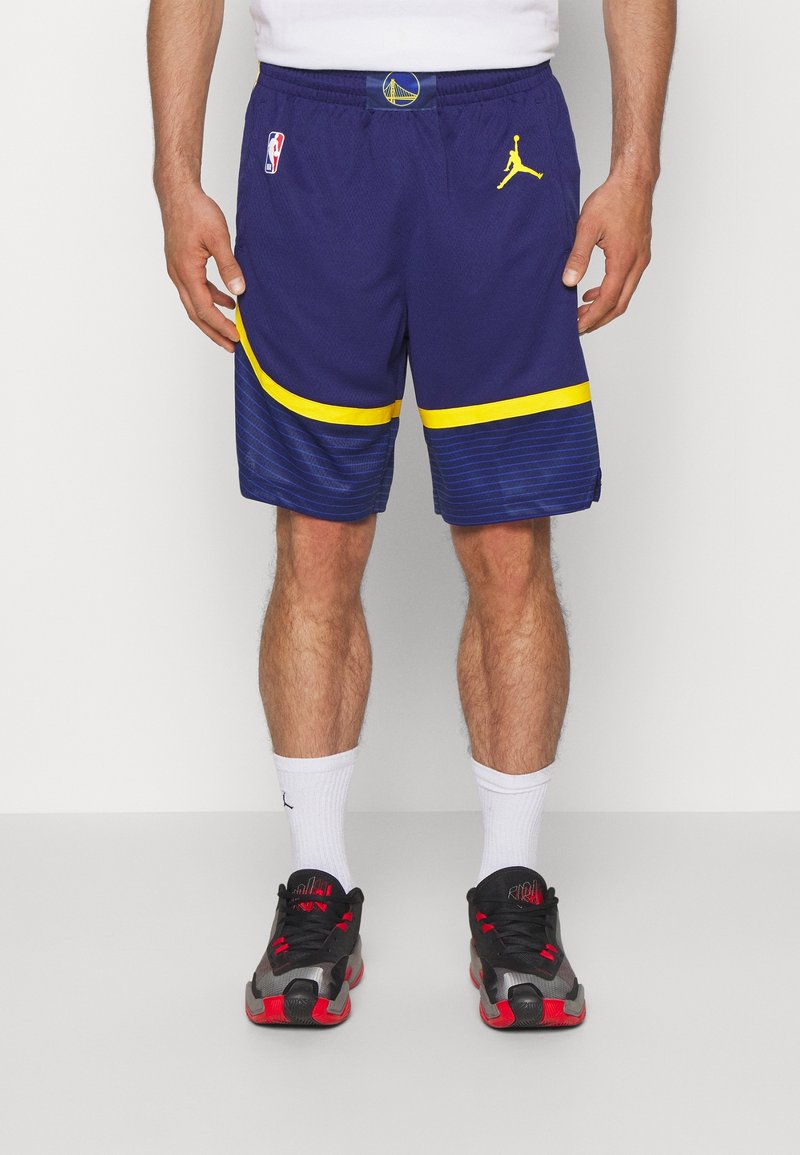 Homme portant un short de basketball bleu et jaune des Golden State Warriors, des chaussettes blanches et des chaussures de sport noires et rouges, debout devant un fond uni.