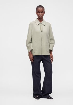 Mannequin féminin portant une veste zippée gris clair avec de grandes poches et un pantalon large foncé, debout sur fond blanc.