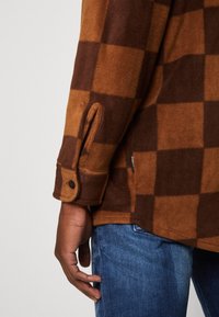 Chemise en flanelle à carreaux marron et orange avec un poignet boutonné, mettant en avant une texture douce et une étiquette latérale partiellement visible. Portée avec un jean en denim.
