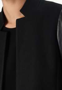Black coat with a high collar, made from textured wool fabric, featuring smooth leather sleeves and a minimalist design.