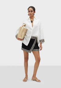 Camicia bianca in lino con vestibilità ampia, abbinata a una gonna nera con bordi a motivo zebra. Gli accessori comprendono una borsa in vimini e gioielli dorati.