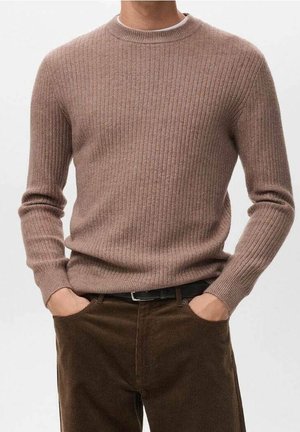 Mężczyzna ubrany w jasnobrązowy, prążkowany sweter z długimi rękawami oraz ciemnobrązowe spodnie sztruksowe, z rękami w kieszeniach.