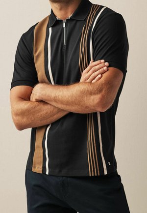Man with arms crossed wearing black polo shirt with vertical brown and white stripes and a zipper collar, dark pants visible.