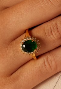 Gold ring featuring an oval green gemstone at the center, surrounded by small white stones, set against a skin-toned background.