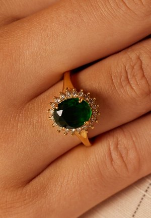Bague en or avec une pierre précieuse verte ovale au centre, entourée de petites pierres blanches, sur un fond couleur peau.