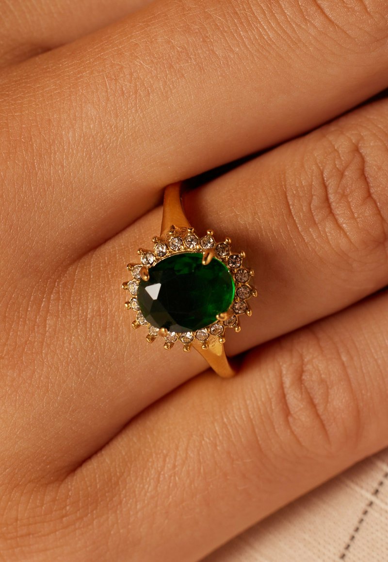 Gold ring featuring an oval green gemstone at the center, surrounded by small white stones, set against a skin-toned background.