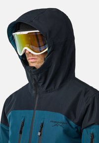 Man wearing black and teal hooded jacket and white ski goggles with reflective amber lenses, zipped up covering neck and chin.