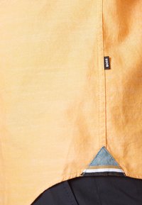 Primer plano del dobladillo de una camisa de tela naranja con una pequeña etiqueta "BOSS" y un parche triangular azul sobre unos pantalones negros.