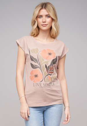 T-shirt rose clair avec un motif floral représentant des fleurs orange et vertes, manches courtes retroussées et col rond. Le texte en dessous indique "VIVEZ SIMPLE ET ÉPANOUISSEZ-VOUS SAUVAGEMENT."