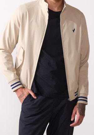 Mann trägt beige Bomberjacke mit blauen Streifen an den Bündchen und einem kleinen bestickten Vogel, schwarzes T-Shirt und schwarze Hose, Hand in der Tasche.