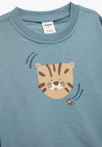 Lichtblauwe sweatshirt met een ronde, beige tijgergezichtafdruk met bruine strepen en snorharen, met de tekst "STAY WILD" eronder. Gemaakt van katoen.