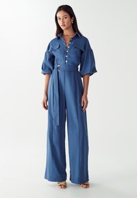 Kvinna som bär en blå jumpsuit med bälte, stora knappar, puffärmar i 3/4-längd och vida ben, stående mot en vit bakgrund.