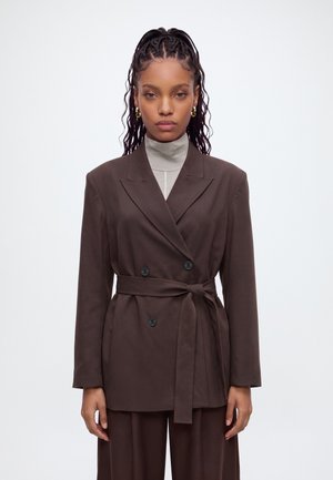 Jeune femme aux longs cheveux tressés porte de grandes boucles d'oreilles en or, un blazer croisé marron ceinturé et un pull à col roulé gris clair.