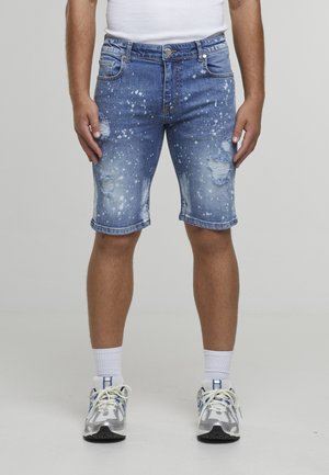 Hombre con zapatillas blancas, calcetines blancos, pantalones cortos vaqueros azules desgastados con efecto de salpicaduras de pintura y camiseta blanca metida por dentro, de pie recto.