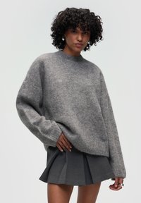 Oversized grauer Pullover aus weichem, strukturiertem Material mit Rundhalsausschnitt und gerippten Bündchen. Kombiniert mit einem dunklen, plissierten Rock.