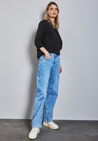 Schwarzes Longsleeve-Oberteil kombiniert mit hellblauen weit geschnittenen Jeans und weißen Sneakern. Das Ensemble hat einen lässigen, entspannten Schnitt.