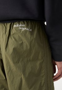 Olivgröna polyesterjoggers med elastisk midja och vit broderad "adidas originals" logotyp nära höfterna.