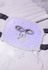 LED ansiktsmask med vit silikonbas och svarta remmar. Har en matris av små lila lampor och ögonavskärningar för behandlingar.