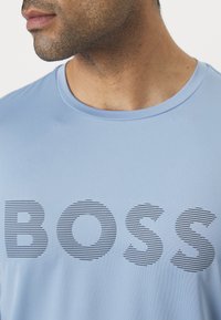 Camiseta de color azul claro hecha de tela suave, con un audaz logo "BOSS" en rayas horizontales negras en la parte delantera. Cuello redondo simple.