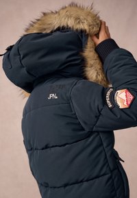 Chaqueta acolchada de color azul marino con capucha con borde de piel, presenta un logo y una insignia cosidos en la manga, puños de canalé y textura acolchada.