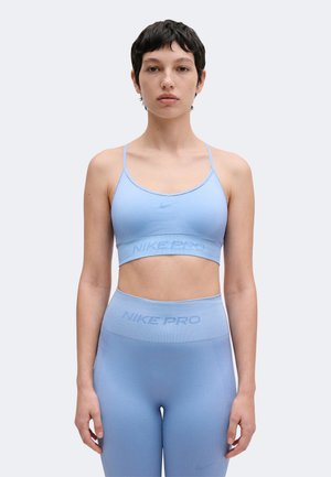 Mujer que lleva un sujetador deportivo Nike Pro azul claro y leggings de cintura alta, de pie frente a un fondo liso y claro.