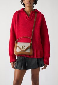 Maglione rosso lavorato a maglia con colletto, abbinato a una borsa a tracolla marrone e beige con un accento dorato. Gonna a quadri con pieghe.