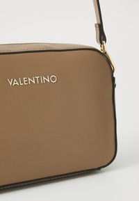 Sac bandoulière en cuir synthétique marron avec logo doré, contour noir, forme rectangulaire, texture lisse et bandoulière réglable.