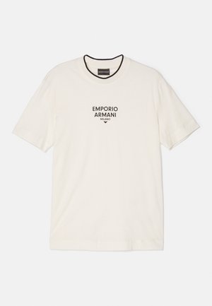 Mies, joka pukeutunut valkoiseen Emporio Armani T-paitaan, mustiin farkkuihin ja mustiin kenkiin, seisoo yksinkertaisen harmaan seinän ja lattian edessä.