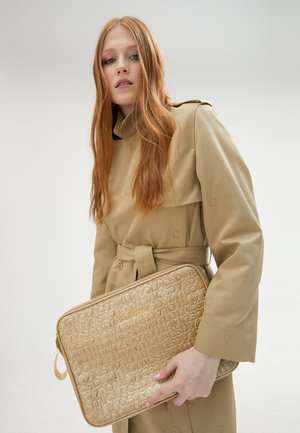 Trench coat beige con cintura e grande patta, abbinato a una borsa per laptop in oro testurizzato impresso con lettere. Superficie liscia e lucida.