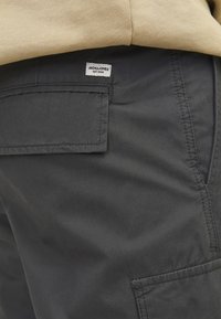 Primo piano di pantaloni grigio scuro con tasca a soffietto e una piccola etichetta bianca con la scritta "Jack & Jones EST. 1990" sotto una camicia beige.