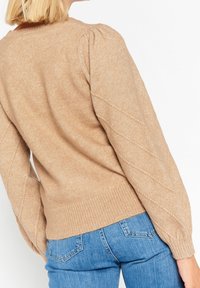 Beige stickad tröja med texturerade, puffiga ärmar och ribbad nederdel. Har ett diamantmönster längs ärmarna. Bärs med blå jeans.