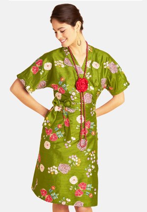 Woman wearing green floral wrap dress with short sleeves and red beaded necklace with large red flower pendant.