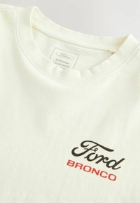 Tricou din bumbac de culoare bej deschis, având logo-ul "Ford" în negru și "BRONCO" în roșu. Guler rotund ribbed și etichetă cosută în guler.