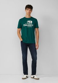 Turbukowy bawełniany t-shirt z białym logo Discovery Channel, w połączeniu z ciemnymi jeansami i białymi trampkami z czarnym paskiem akcentującym.